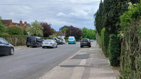 A street in Northampton, with several cars on it, parked and being driven. A path is to the right, with patches on it, with bushes and trees down the right-hand side. There are houses to the left, with more trees and bushes and more parked cars.