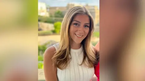 Hannah Dyos smiles at the camera wearing a white dress, she has long blonde hair.