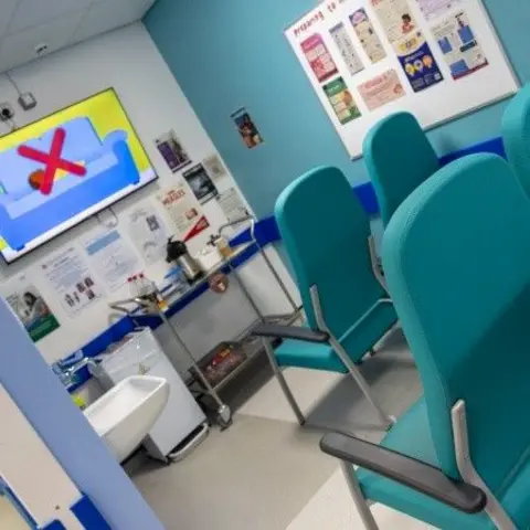 NHS Humber Health Partnership A waiting or consultation room in a hospital with teal padded chairs arranged along the right side. The room has light blue walls and a whiteboard on the right wall displaying various posters and information leaflets. On the left, a wall-mounted digital screen shows a graphic with a large red “X” over a sofa. Below the screen, a counter holds medical supplies, including gloves, containers, and small bottles. A small sink with a tap is positioned beneath the counter on the left.