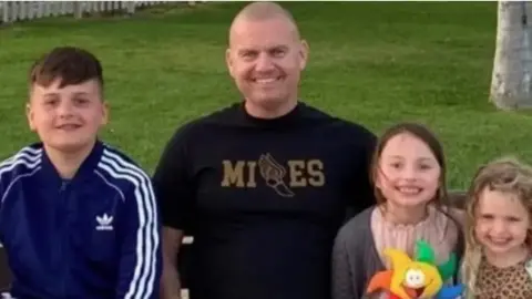 Handout A smiling Dean Miles pictured with his three children - a young boy with brown hair and two girls, one with brown hair and the other with blonde hair. They are also smiling in the photo.