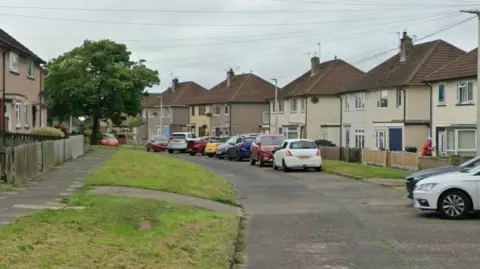 Whinsmoor Drive in Carlisle. Beige semi-detached houses line either side of a curving road which has grass verges and parked cars down one side.
