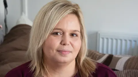 A young woman with shoulder length blonde hair, wearing a burgundy top and sitting on a brown sofa.