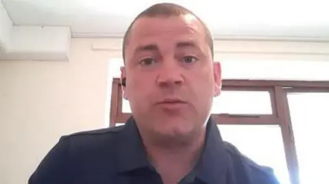 Colin Nulty with short brown hair wearing a blue shirt sitting in a room in a house with a window in the background.