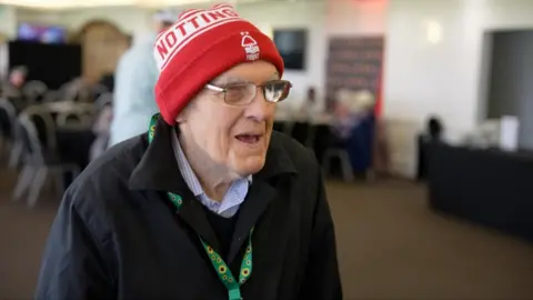Frank Wignall at the dementia support group Forget Me Notts in a function room at Nottinghamshire Cricket Club. He is smiling wearing a modern day Nottingham Forest hat with the club logo proudly displayed 