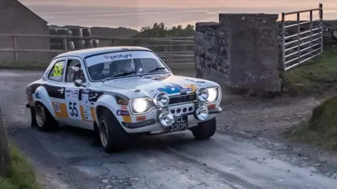 LUCAS CROYDON PHOTOGRAPHY A white rally car with it headlights on going past farm gates. It has the number 55 on its side.