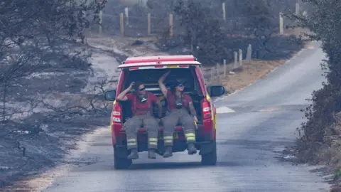 SFRS Two fire officers in the back of a fire service vehicle. The boot door is open and they are sitting in the back with their legs dangling from the vehicle. They are covered in smoke and wear sunglasses. The vehicle is moving down a road and either side is the blackened remains of trees and shrubbery.