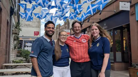 Kings Four people - two men and two women - stand in the street outside a coffee shop, with an array of small Scotland flags hung up across the street. The group are all wearing either Scotland tops or dark blue shirts.