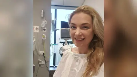 Gemma Bailey Gemma is pictured in a hospital gown whilst recieving cancer treatment. She has long blonde hair and is smiling at the camera.