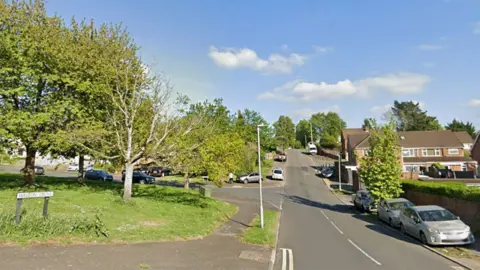 The turning for Walton Drive, with a street name sign on the left that is on top of a grassy area with trees. On the right are parked cars on a verge, a tree and some housing. The road goes up a slight incline into the distance.