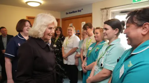 Queen Camilla wearing a black coat while meeting hospice nurses who are wearing a teal uniform. They are stood in a hall near wooden doors.