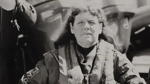 Jack Lowe (Lifeboat Station Project) A black-and-white photo of Liz staring straight ahead and standing in front of an RNLI vessel. 