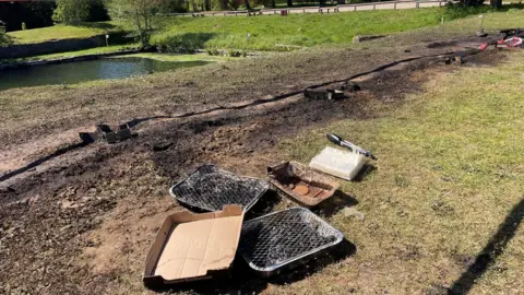 Burnt out disposable BBQs are on the floor, surrounded by fire-damaged grass. In the background is a lake and grassland.
