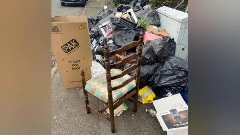 Steven Message A pile of black bin bags on a pavement, with some split open and rubbish on the floor. There are cardboard boxes on the pavement, and a wooden chair as well.