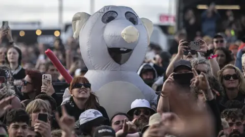 Getty Images A person in a large, white rat costume stands in the middle of a crowd of people, many of whom are holding up their mobile phones