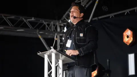 Getty Images Dom Joly has dark hair and is standing at a plinth on stage. He is in a black denim jacket with dark tshirt and jeans. His mouth is open as he speaks into a microphone headset