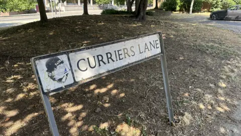 A road sign at the edge of a residential street. The sign reads Curriers Lane. There are trees behind the sign and a grass area. A lone car is parked further up toward the right. 
