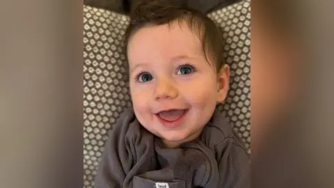 Family photograph Eight-month-old Rupert with brown hair wearing a cream top and brown knitted braces. He is smiling.