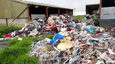 The image shows large piles of assorted waste scattered across the outdoor area of a farm. Two buildings with corrugated metal roofs and partially open sides are visible in the background.
