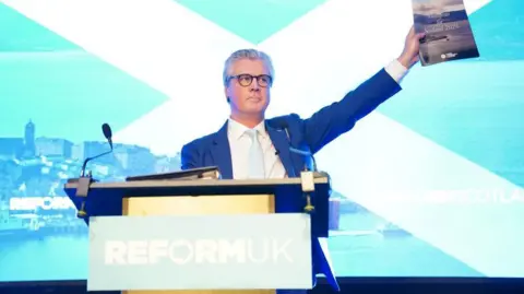 PA Media A politician wearing a deep blue suit holding up a manifesto in front of a saltire projected on a screen behind