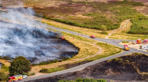 Lewis Johnstone Photography Fire engines parked on roads around a heath which is black from fire, pockets of flames can be seen and large amounts of smoke.