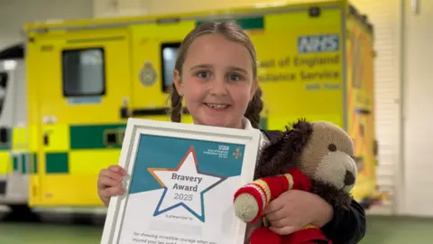 Bunny-Mae who is standing in front of an ambulance with her hair in two plaited pig tails. She is looking directly at the camera and smiling. She is holding a bravery award certificate and a teddy bear.