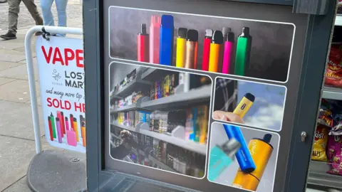 The lower half of a shop door with pictures of brightly-coloured vapes on it. Just outside the door is a sign that reads in red letting 'Vape - Lost Mary and many more top brands sold here' on a white background.