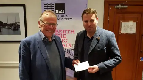 Hull Maritime Two men smile and hold a cheque at a presentation – they are both wearing suits and standing in front of a Hull Maritime project promotional banner.