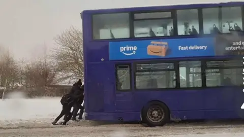 Fubar News A group of people dressed in black push a double decker bus on a snowy road