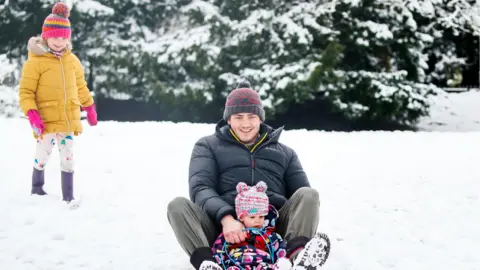 PA Media Dan Ives with his daughters Eden (on the sledge) and Poppy (left) in a snow covered Buxton Park in Buxton, Derbyshire