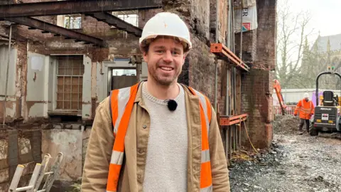 Person wearing a hard hat and high-visibility vest standing inside the stripped-out Royal Clarence redevelopment site, with exposed brick walls, steel supports and construction work underway in the background.