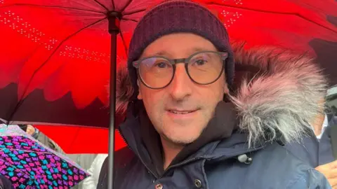 Robert Rosenfield is standing under a red umbrella during the vigil. He is wearing a navy hat and coat and glasses. 
