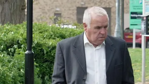 Kevin Miller is wearing a grey or navy blue pinstripe suit while on a pavement or footpath. There are bushes and signs behind him.
