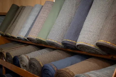 Kirsty Anderson/ Lovat Mill About a dozen rolls of tweed fabric in various colours. They are sitting on two shelves. 