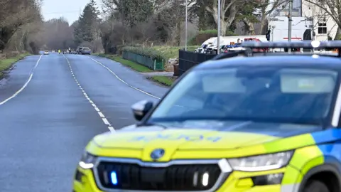 Pacemaker A police car, blurred, is parked across a main road. There are rows of trees and on either side of the road and a white building on the right. 