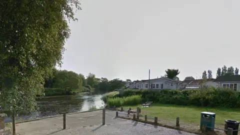 Wooden posts along river bank with tree lined river off to right and number of park homes alongside