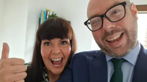 Halina Rifai A woman with brown hair and a fringe, she is making a thumbs up gesture and has a wide smile expression. She is posing with a bearded man wearing glasses who is also smiling.