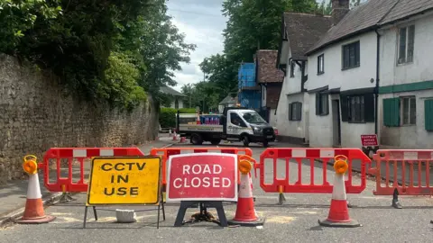 Surrey Highways A red sign reading 'road closed' and yellow sign labelled 'CCTV in use' sit in front of temporary orange fences and cones, closing off part of the A281, also known as 'The Street'. Scaffolding has been erected to fix a cottage in the background behind the fencing.