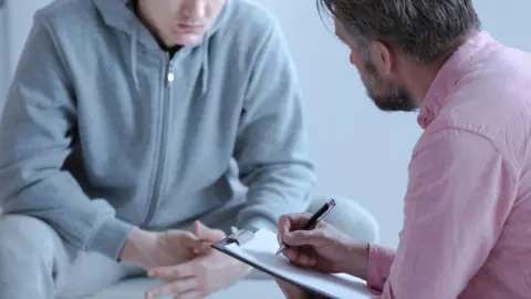 A stock photo of a psychologist who is wearing a pink shirt interviewing his patient who is in a grey tracksuit during a therapy Counselling Session