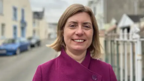 Lyndsay Feltham - a woman with a brown bob wearing a purple coat smiling at the camera. The background of a main road is blurred.