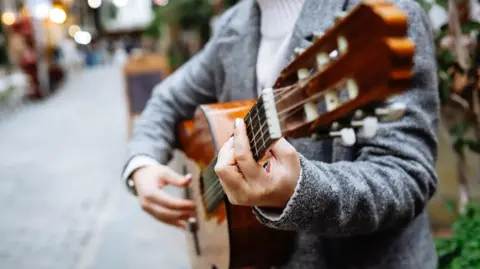 Generic image of a woman playing acoustic guitar by the side of a pedestrianised street with a blurred background of street cafe seating and buildings with lights showing. She is wearing a grey jacket and white polo-neck. Her face is not visible.