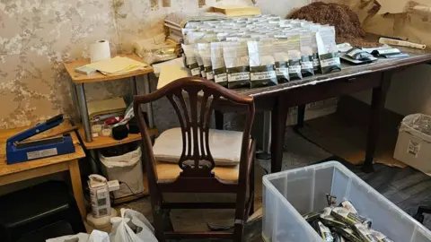 On an old-fashioned table there are rows upon rows of bags of tobacco, as well as boxes on the floor