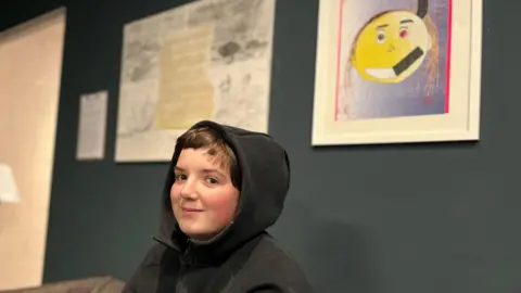 Federica Bedendo/BBC Leo sat under his artwork. Leo has short brown hair and is wearing a black hoodie with his hood pulled up over his head. His drawing shows a yellow emoji-like face with dark brown hair, a black horn, black smile and a red eye on one side. The other side has a brown eye and a white smile. 