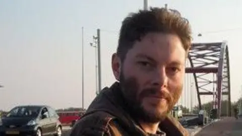 Family handout James Self has brown hair, a beard and is wearing a brown jacket. He is standing by a bridge and smiling.