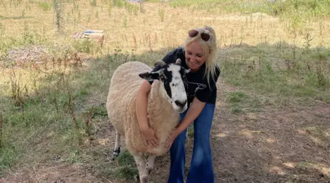 Charity founder Hayley Prescott, who has blonde hair in a ponytail and is wearing jeans and a black t-shirt, hugs a very large sheep. He has black and white markings on his face and small horns. He reaches Hayley's hip.