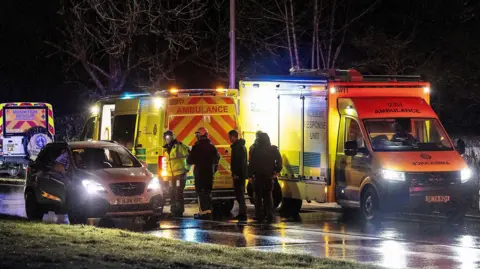 Ambulance vehicles and a mountain rescue vehicle parked on a road. A car is parked next to the emergency vehicles. It is dark. A group of people are stood next to the vehicles.
