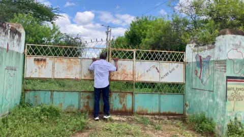 Wycliffe Muia / BBC A man wearing a white shirt and black trouser stands at a large weathered metal gate of closed Sooma Primary School, peering through it into a green compound surrounded by trees