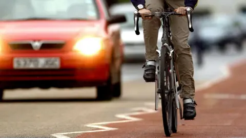 A cyclist on a red cycle lane in Cambridge. All that can be seen is the cyclist's legs and their hands holding a dropped handlebar. On the left is a red car with its lights on and behind it are more cars, all blurred out.