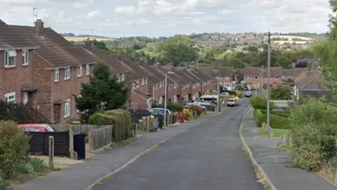 Google A mid-20th century residential street runs downhill with red-brick, semi-detached houses to either side and more at the the bottom of the hill. Cars are parked in driveways and some by the side of the road. In the background hills rise from a valley with more homes visible in the distance. 