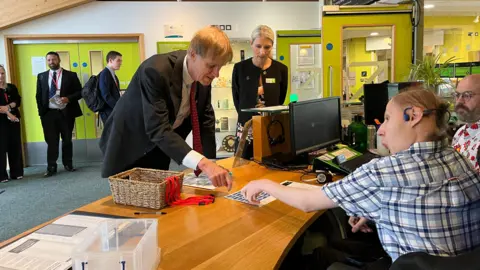 Everyone is in the reception area of the National Star College. Minister Stephen Timms is leaning over a counter, speaking with student Sam seated behind it. The counter is cluttered with typical office items like computers, papers, and supplies. They are pointing at the inclusive disability badges. In the background, a few other people are standing and observing the interaction.