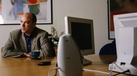 Getty Images Man at a desk with computers and pictures on the wall